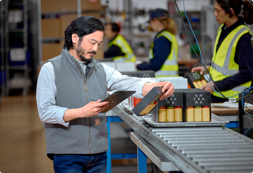 A Shopify merchant with dark hair and facial hair is holding a tablet while standing next to packaging equipment in a warehouse fulfillment center. He is wearing a light gray zip-up jacket over a collared shirt and jeans. In the background, workers in high-visibility safety vests are operating in the warehouse environment.