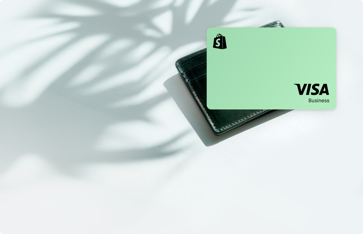 A mint green Shopify Credit card sits on top of a black leather wallet on a white surface with plant shadows. The card features the black Shopify logo in the upper left corner and "VISA Business" branding in the lower right corner.