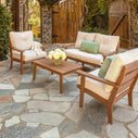 stone patio with wood outdoor loveseat and two chairs all with cream cushions and sage accent pillows, matching wooden outdoor coffee table with vase with yellow flowers in it.